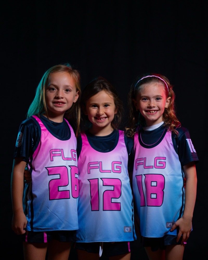 Three young FLG players in pink and navy pinnies, smiling at the camera.